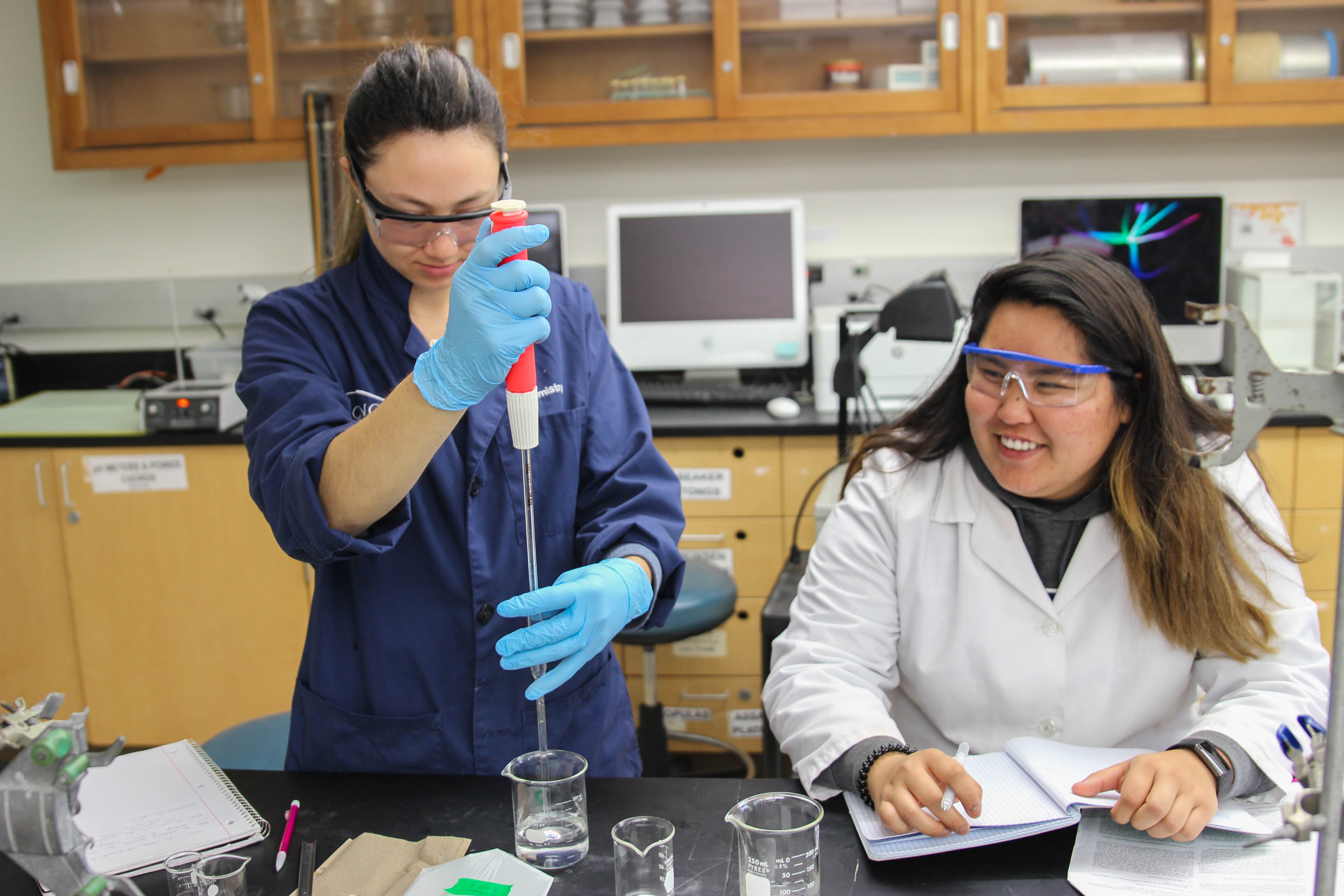 A chemistry student in a blue lab coat uses a pipette while another chemistry student takes notes