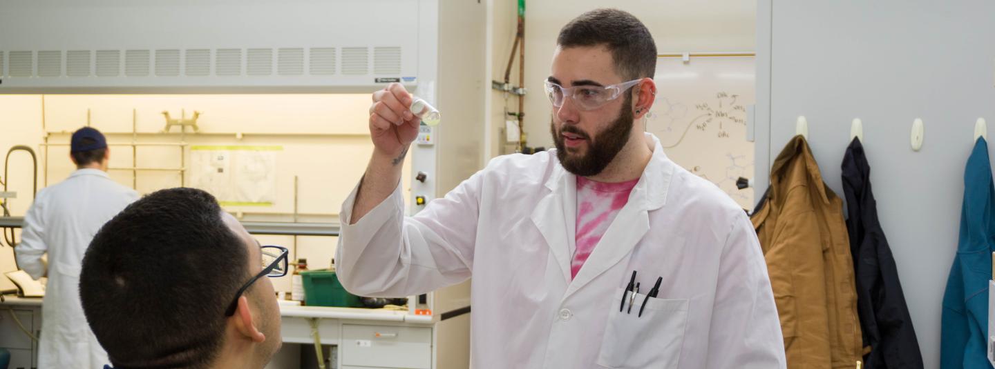 A student in lab coat and protective eyeware examines contents of a vial