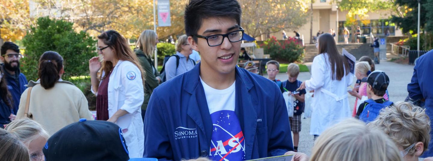Student in blue labcoat and NASA shirt talks with a group of children