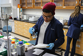 A student in a blue lab coat holds an Erlenmeyer flask in one gloved hand while looking at a lab notebook in his other