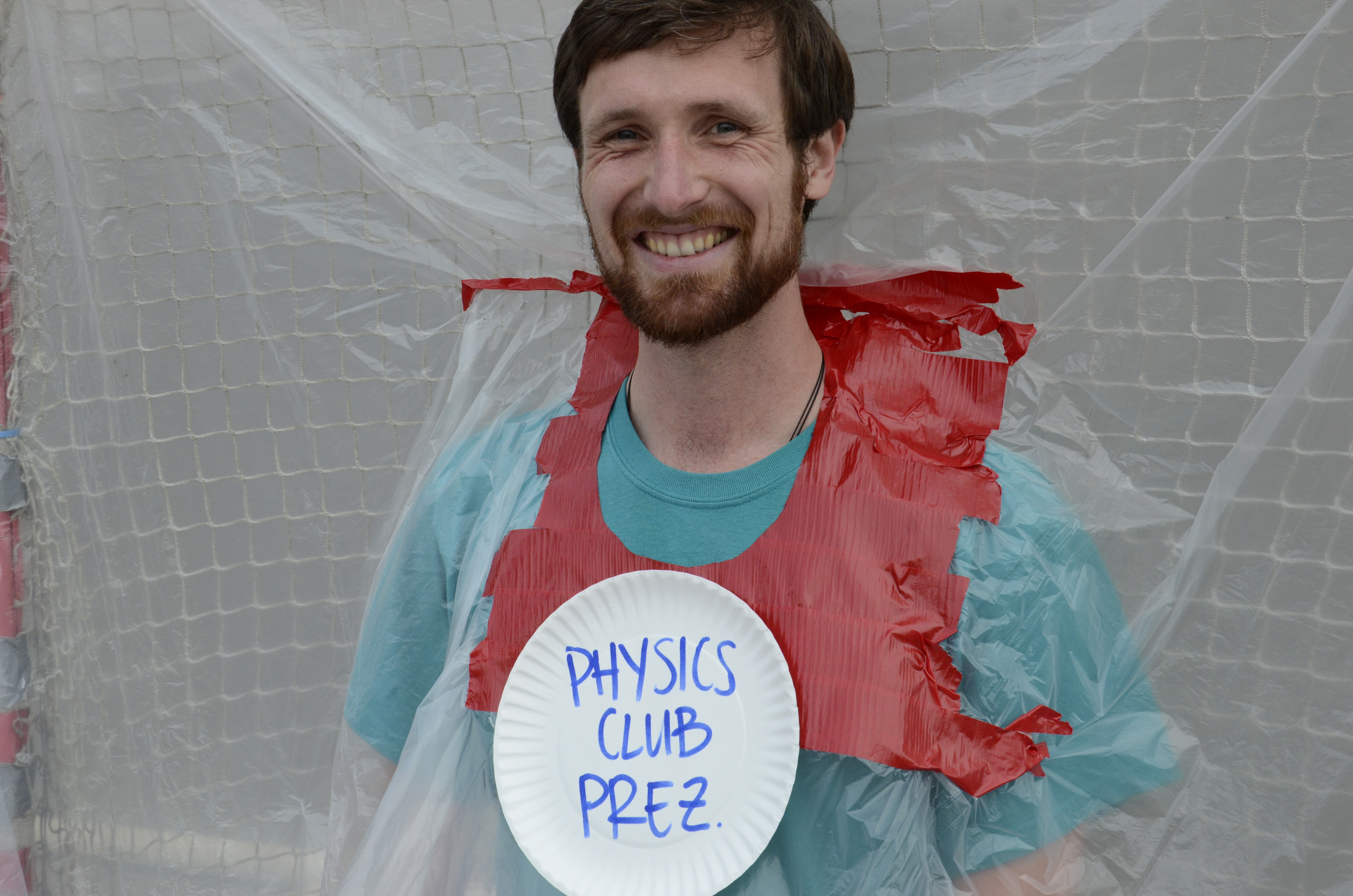 Physics club president sits with his head sticking out out of a plastic sheet for Pi Day and wearing a sign that says "Physics Club Prez"