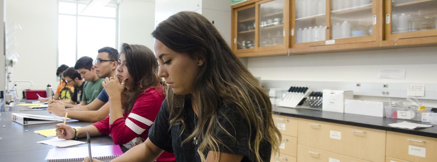 Students sit at a lab bench taking notes with glassware in cupboards in the background