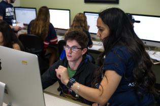 A student helps another at a computer with more computers and students in the background in a computer science lab