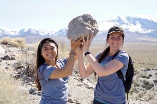 Two students hold a large rock above their heads in a dessert with snow capped mountains in the background